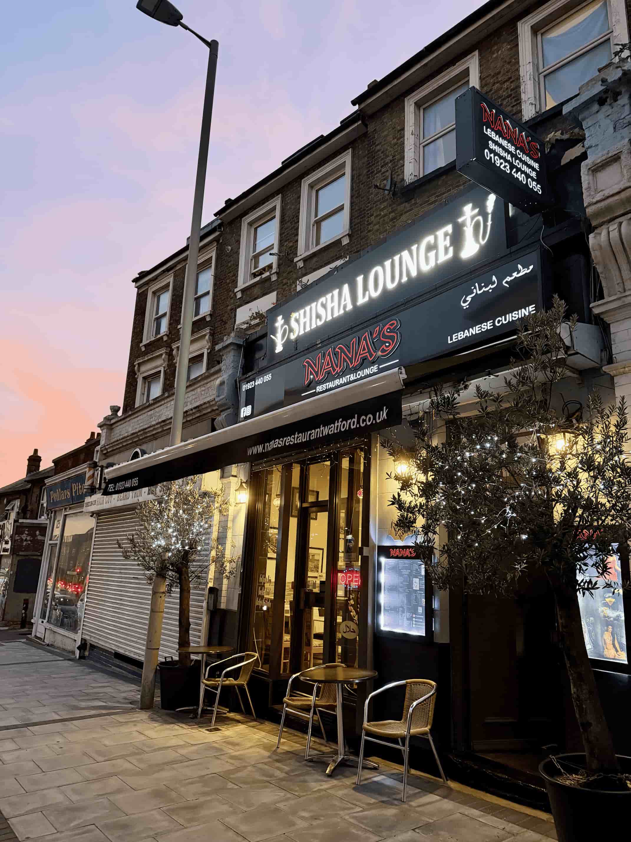 Nana's shopfront at dusk: the lit Shisha Lounge and Lebanese Cuisine signs, fairy-lit olive trees and a table and chairs on the pavement